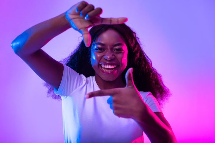 Capturing moments. Happy black woman making picture frame with fingers, looking at camera and smiling for photo in neon light. African American lady showing photograph gesture